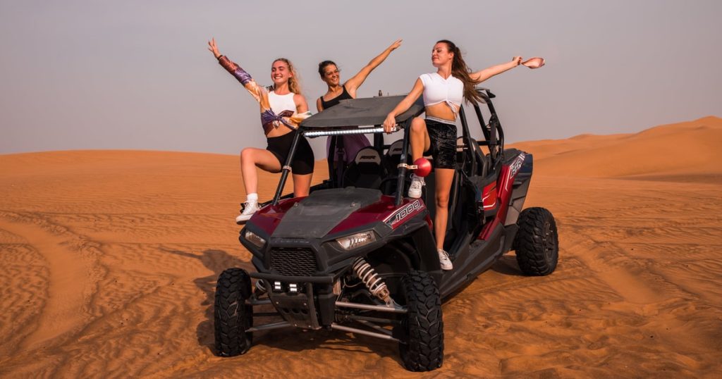 Two travelers in a2 Hour Dune Ride Dubai Can-Am Maverick buggy, wearing helmets and goggles, laughing while driving through the Dubai desert.